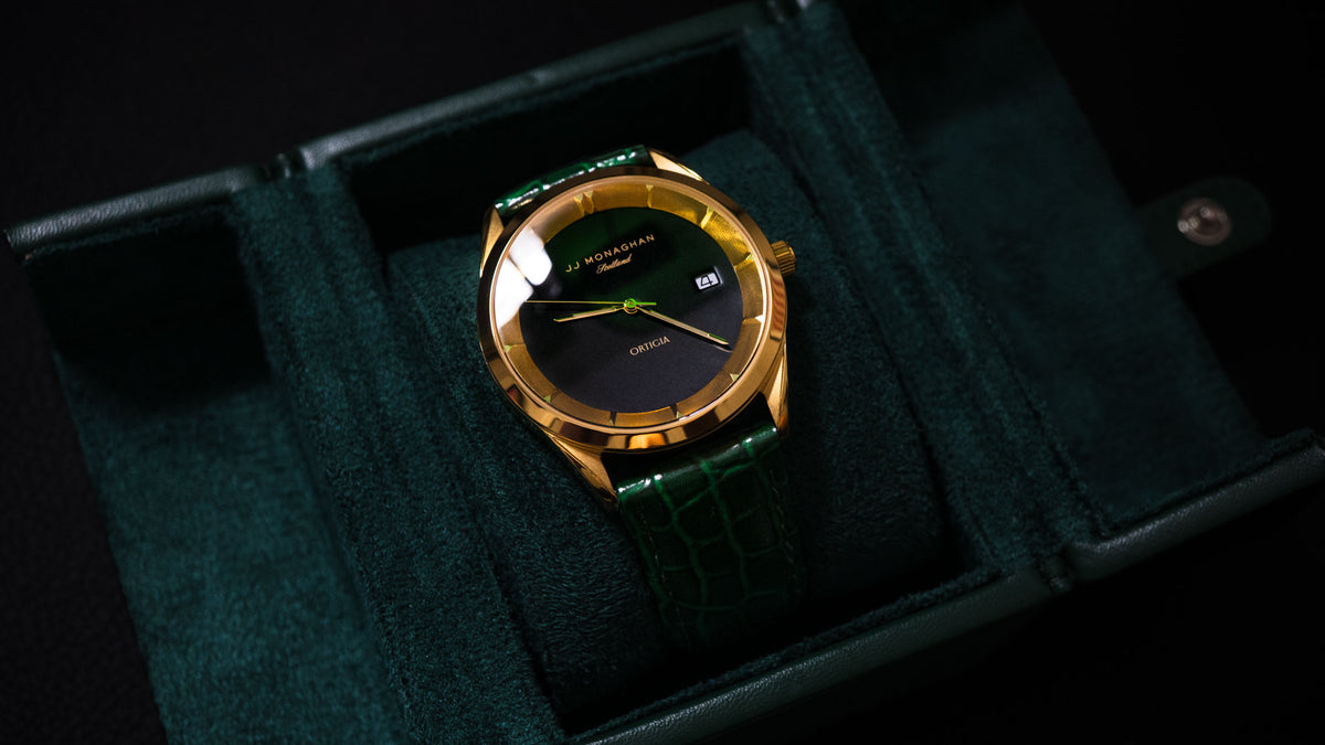 The Ortigia, a gold-cased watch with a black dial and dark green leather strap, resting in its green watch box