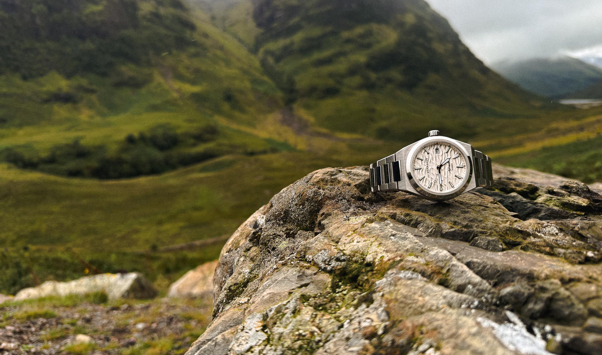 A JJ Monaghan watch resting on a rock above a green mountain valley